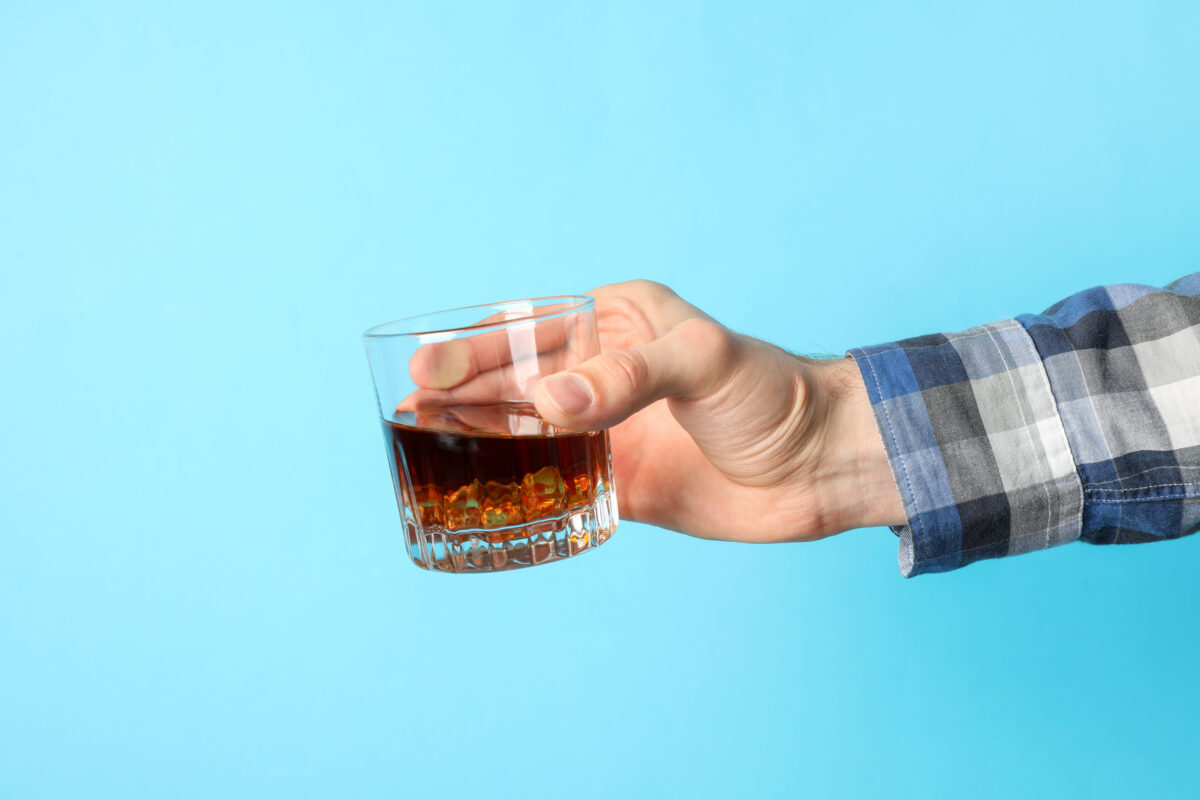 Foods You Should Never Eat Before Bed 3 Male hand holds glass of whiskey with ice cubes on blue background, space for text
