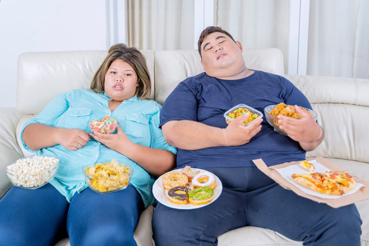 7 Surprising and Often Overlooked Ways Greasy Foods Can Harm Your Body and Affect Your Health 1 Portrait of fat Asian couple sleeping, while watching television with boredom and junk foods scattered around them in their living room
