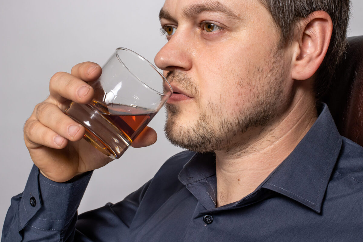9 Foods That May Be Sabotaging Healthy Aging 4 A handsome young man of 30-35 years old in a shirt sits on a brown leather armchair and holds a glass of whiskey in his hand.