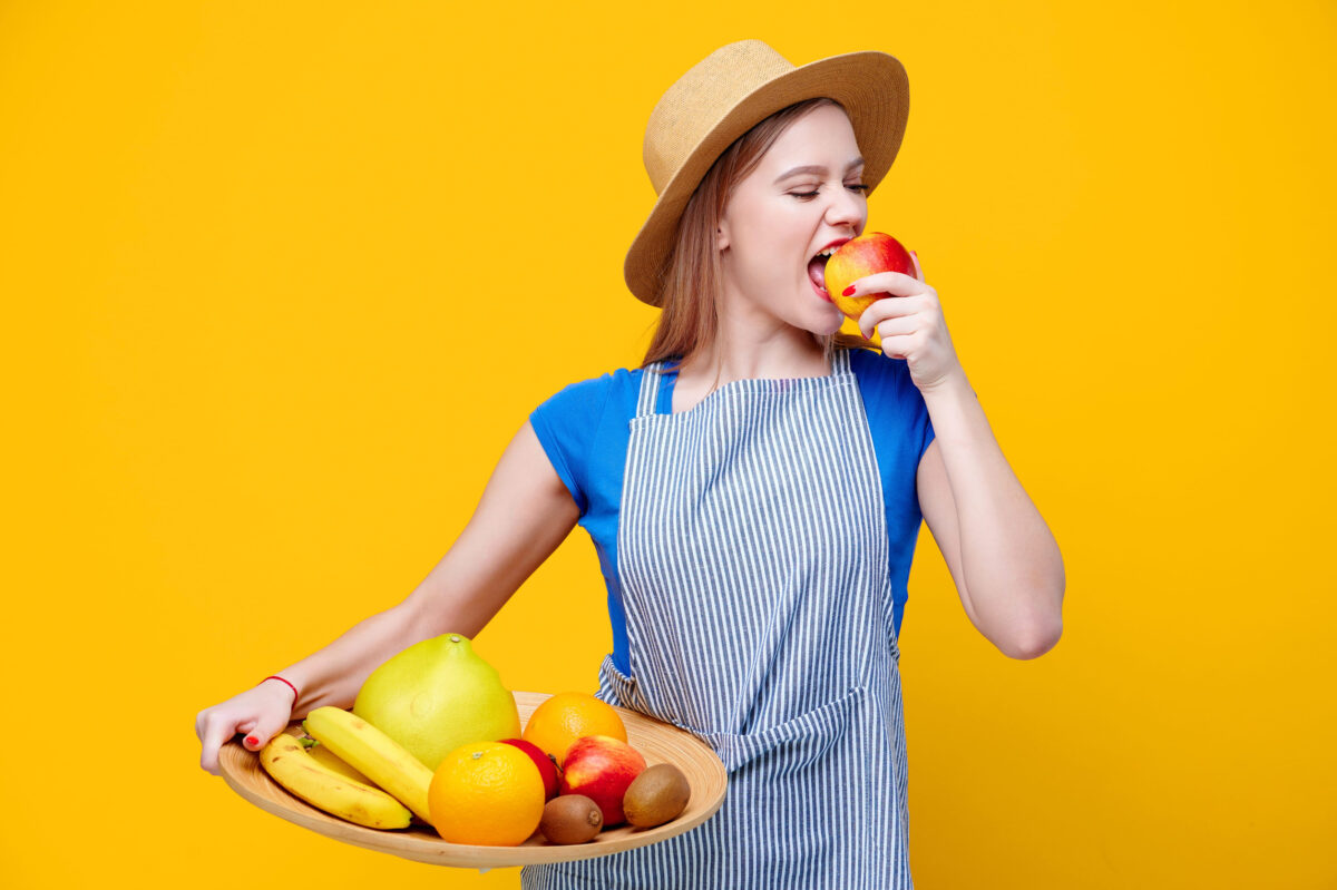 The Hidden Dangers of Juice 4 Caucasian female gardener holding fruits on wooden plate eat apple dressed in straw hat and an apron on yellow background