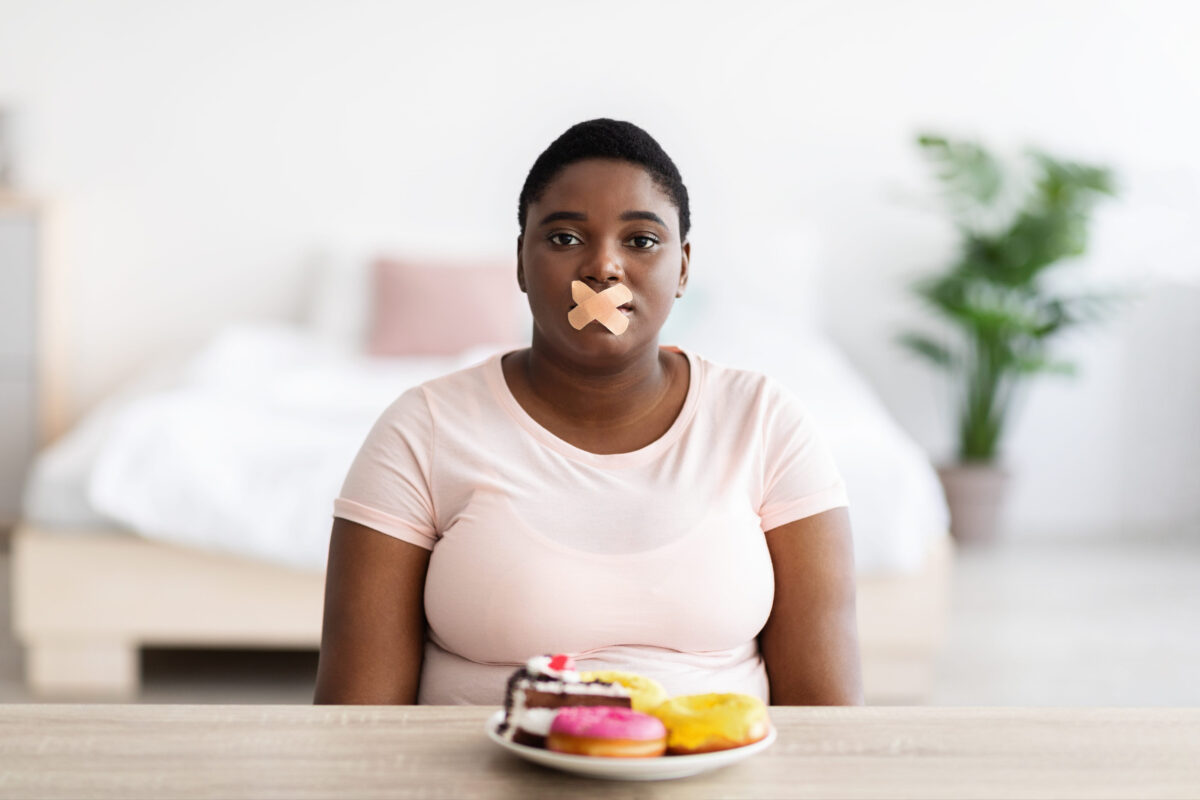 8 Impact of Long-Term Keto Diet on Your Health 1 Upset curvy black woman with bandage on mouth sitting at table with plate of sweets, refusing desserts, losing weight
