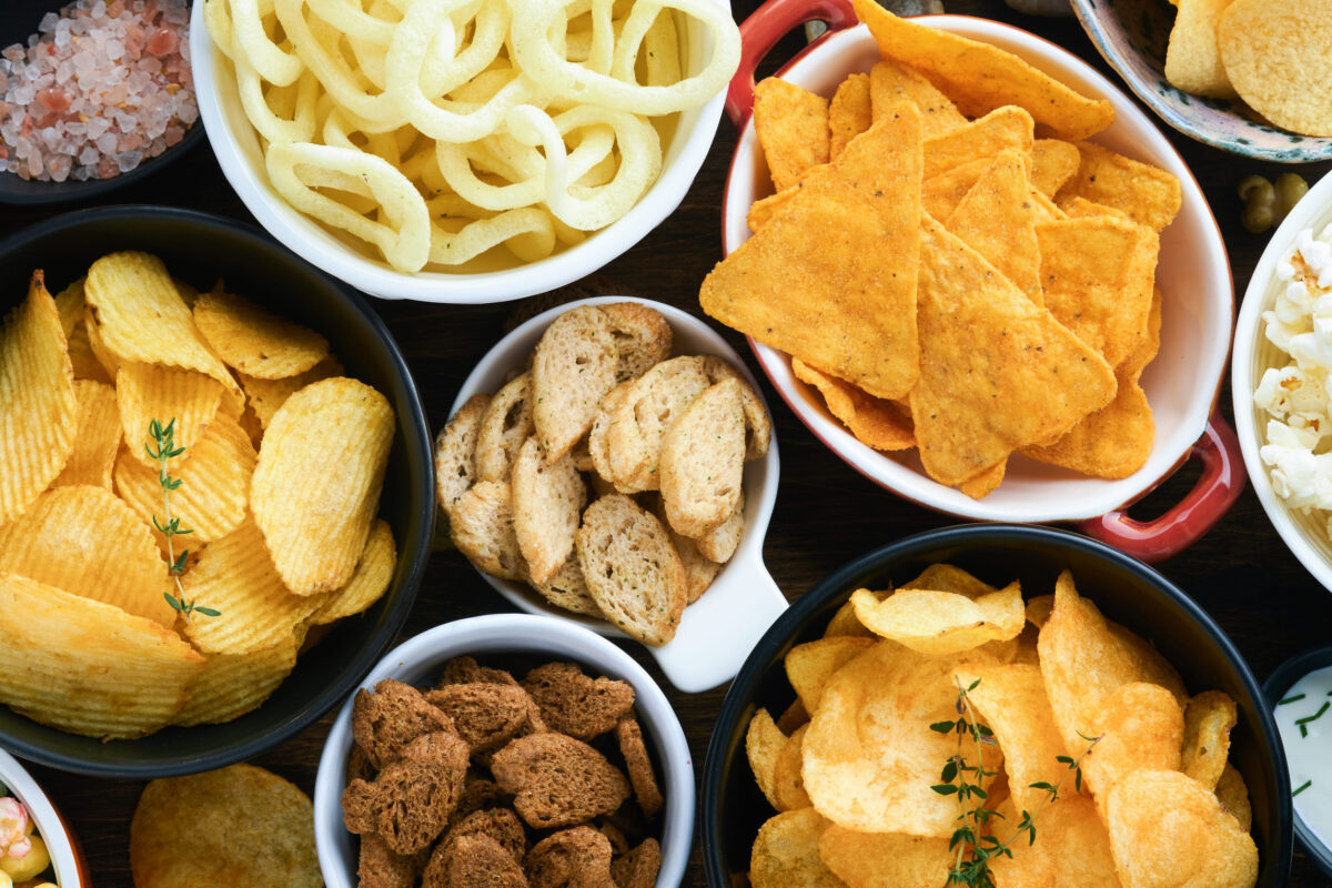 The 6 Most Common Foods That Could Be Causing Your Food Cravings 2 Unhealthy foods or snacks. All classic potato snacks with peanuts, popcorn and onion rings and salted pretzels in bowl plates on old wooden background. Unhealthy food for figure, heart, skin, teeth.