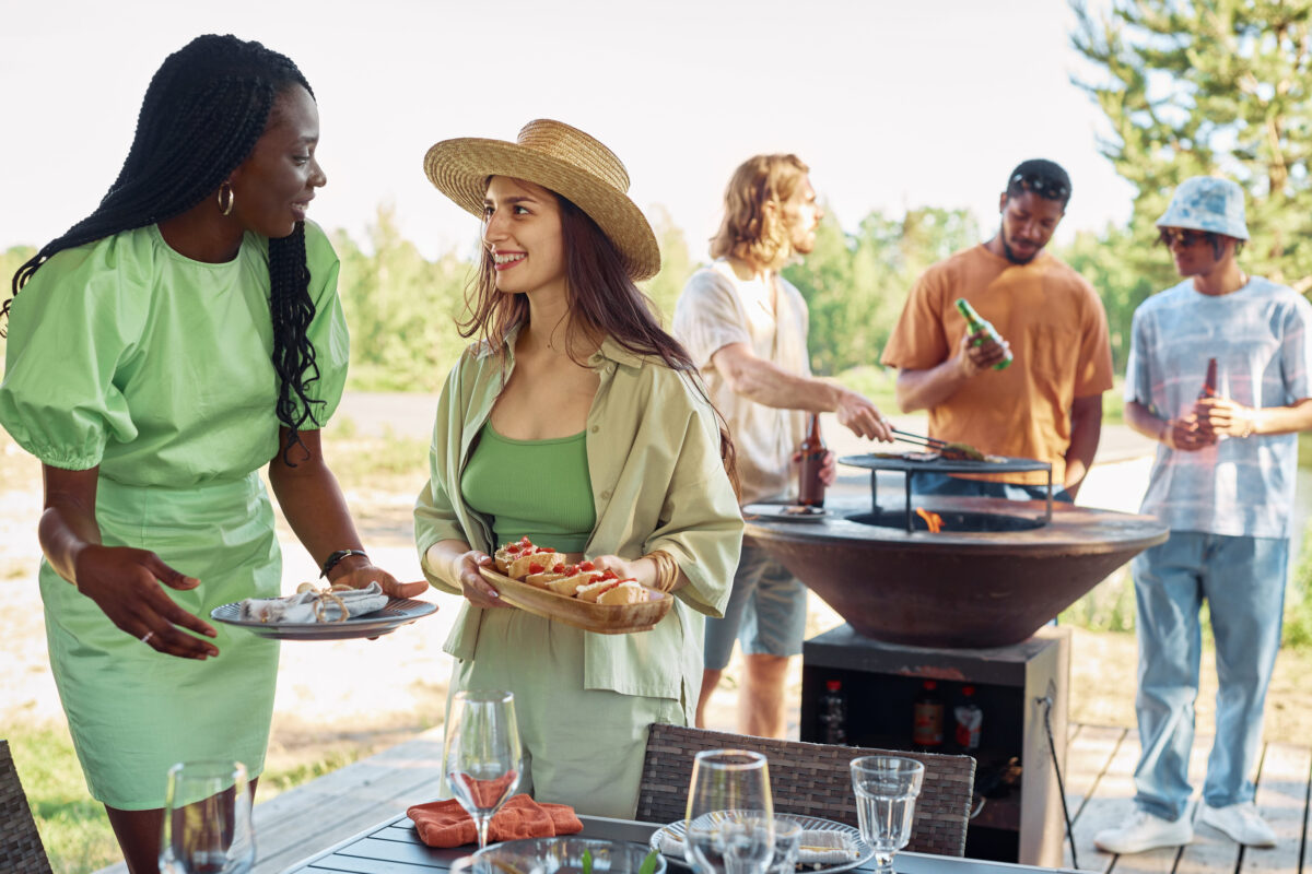 8 Etiquette Mistakes That Will Make You the Worst Guest at a Barbecue 1 Diverse group of young people enjoying barbeque party outdoors in Sumer, focus on two smiling women serving food, copy space