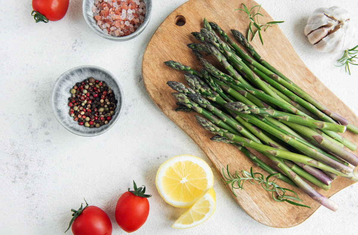 10 Vegetables You Won’t Find on My Plate—And Why I Skip Them Every Time 4 A bundle of fresh asparagus is laid out on a wooden cutting board alongside sliced lemons, robust tomatoes, garlic, and a mix of seasoning. The arrangement suggests the preparation of a healthy, flavorful meal, with the ingredients fresh and prominently displayed against a light background.