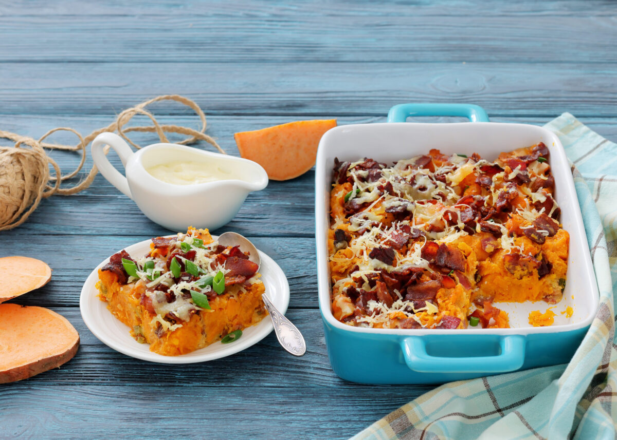6 Thanksgiving Dishes That Will Shock Your Guests 4 savory sweet potato casserole with crispy fried bacon, cheese, green onion in blue baking dish on wood table with ingredients, horizontal view from above, close-up, not AI generated