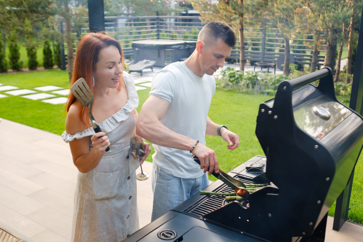 8 Etiquette Mistakes That Will Make You the Worst Guest at a Barbecue 3 Two friends stand in a backyard while grilling food on a barbecue. One holds a spatula and the other is using tongs. They enjoy drinks and share laughter in a garden setting.