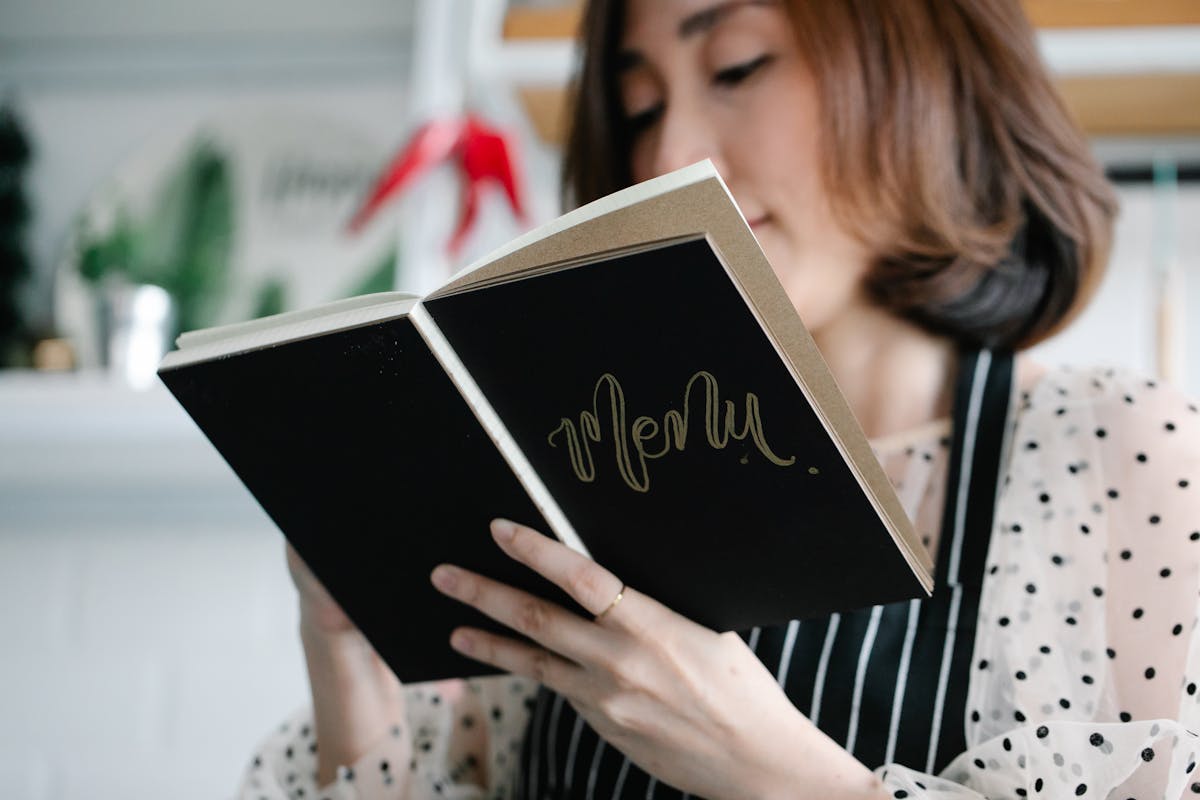 10 Grocery Tips To Avoid Ruining Your Budget 1 A chef in a polka dot blouse holds a menu book in a stylish restaurant, ready to present the dishes.