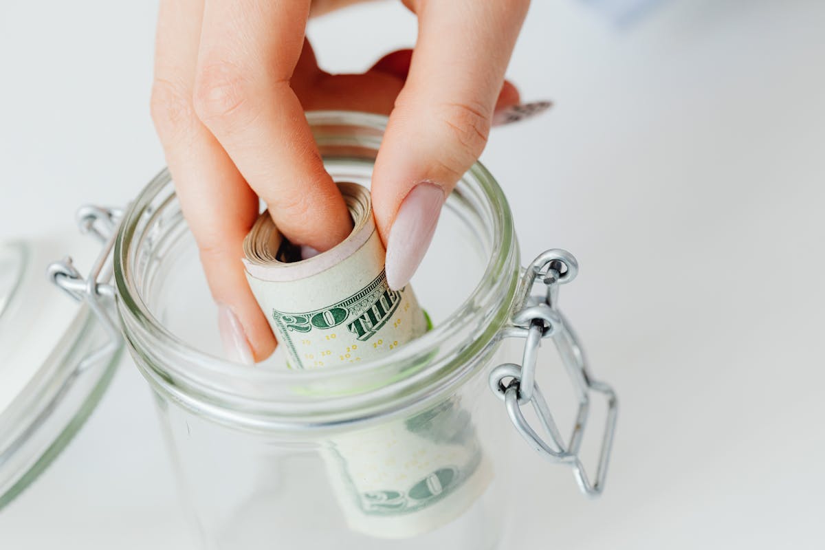 Ignoring These 6 Financial Mistakes Could Cost You Everything 1 A close-up of a hand placing rolled dollars into a glass jar, symbolizing savings.