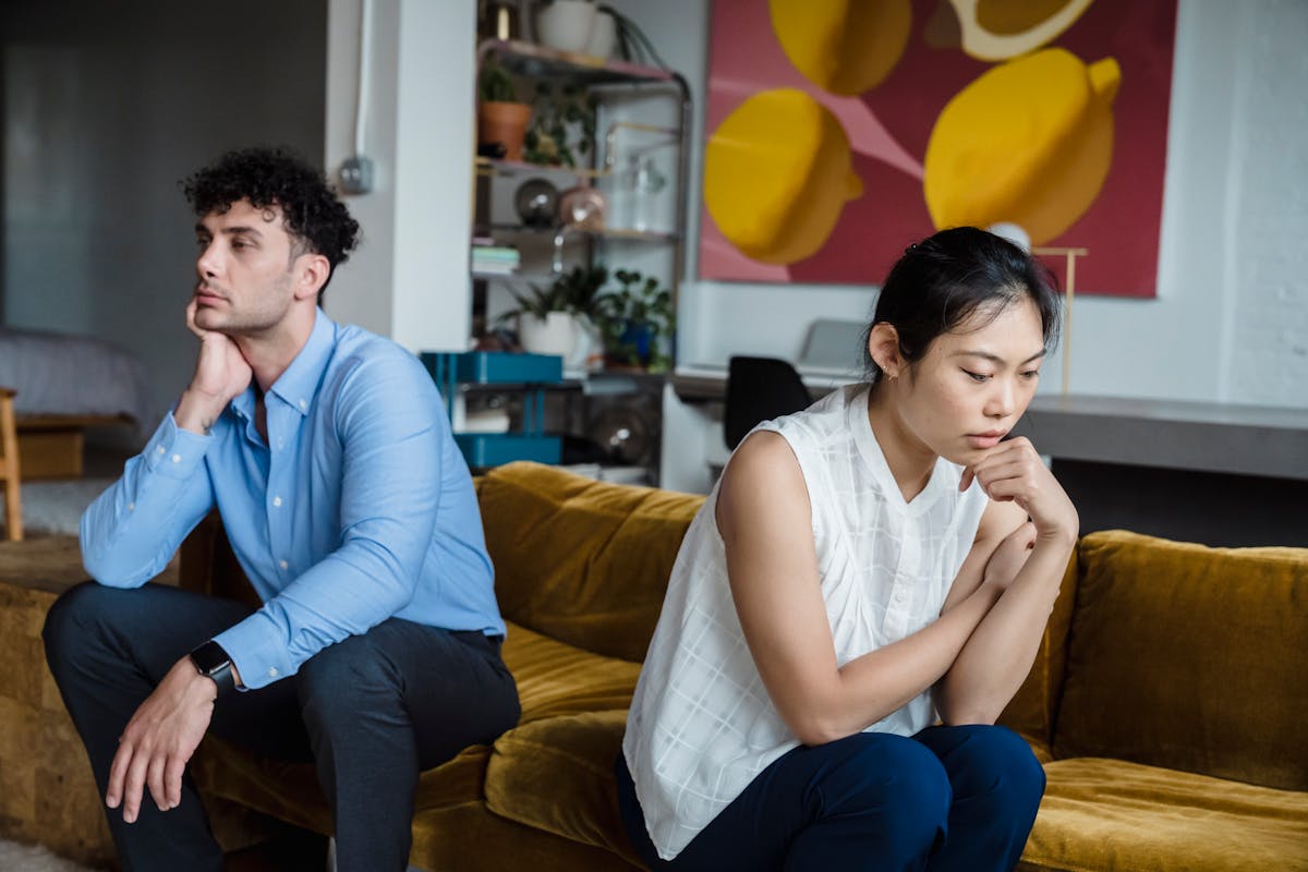 8 Relationship Habits That Are Slowly Killing Your Bond 2 A couple sits indoors on a sofa, both appearing thoughtful and contemplative.