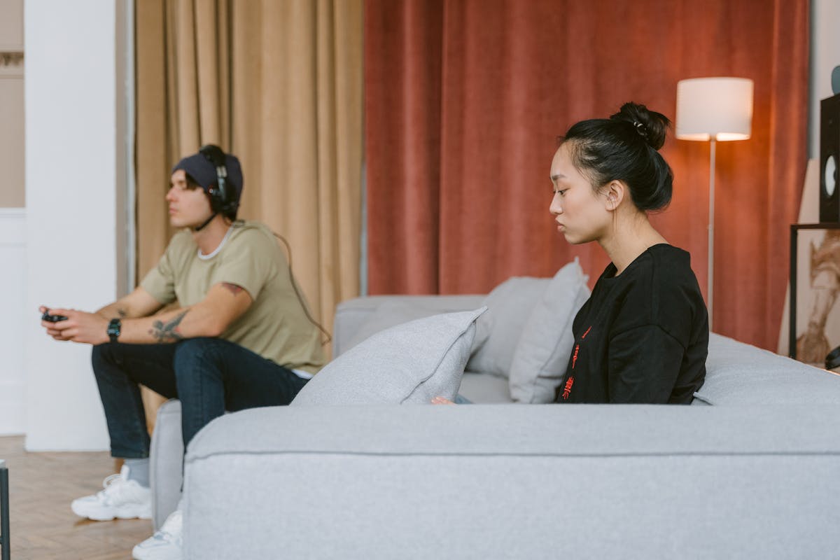 8 Everyday Mistakes That Slowly Undermine Your Marriage 3 A couple sits on a couch, illustrating tension as one plays video games and the other appears distant.