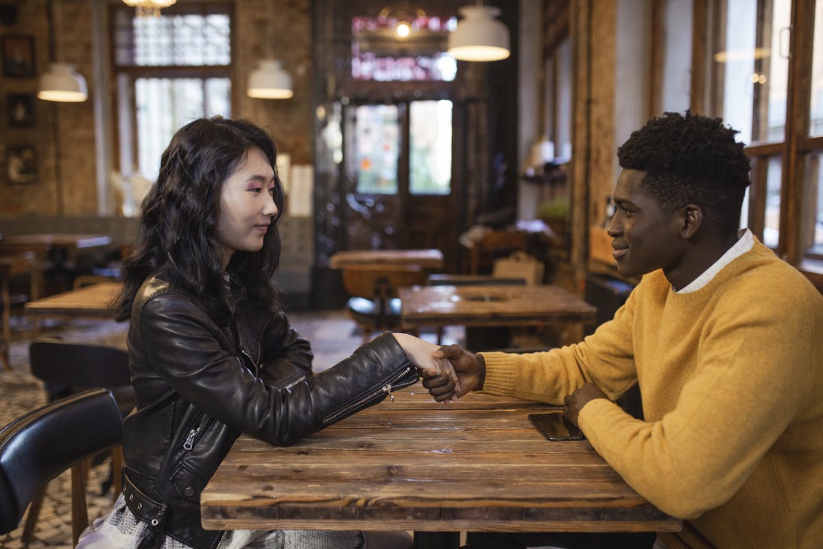These 8 Silly Fights Are Ruining Your Relationship Without You Knowing 4 A diverse man and woman sit in a café, shaking hands and smiling warmly.