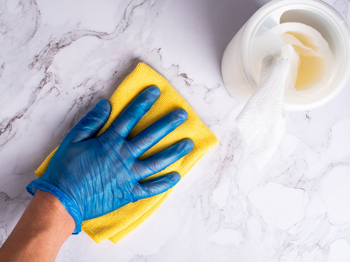 15 Cleaning Products You Should Avoid at All Costs and What to Use Instead 2 A gloved hand cleans a marble countertop with a yellow cloth next to disinfectant wipes.