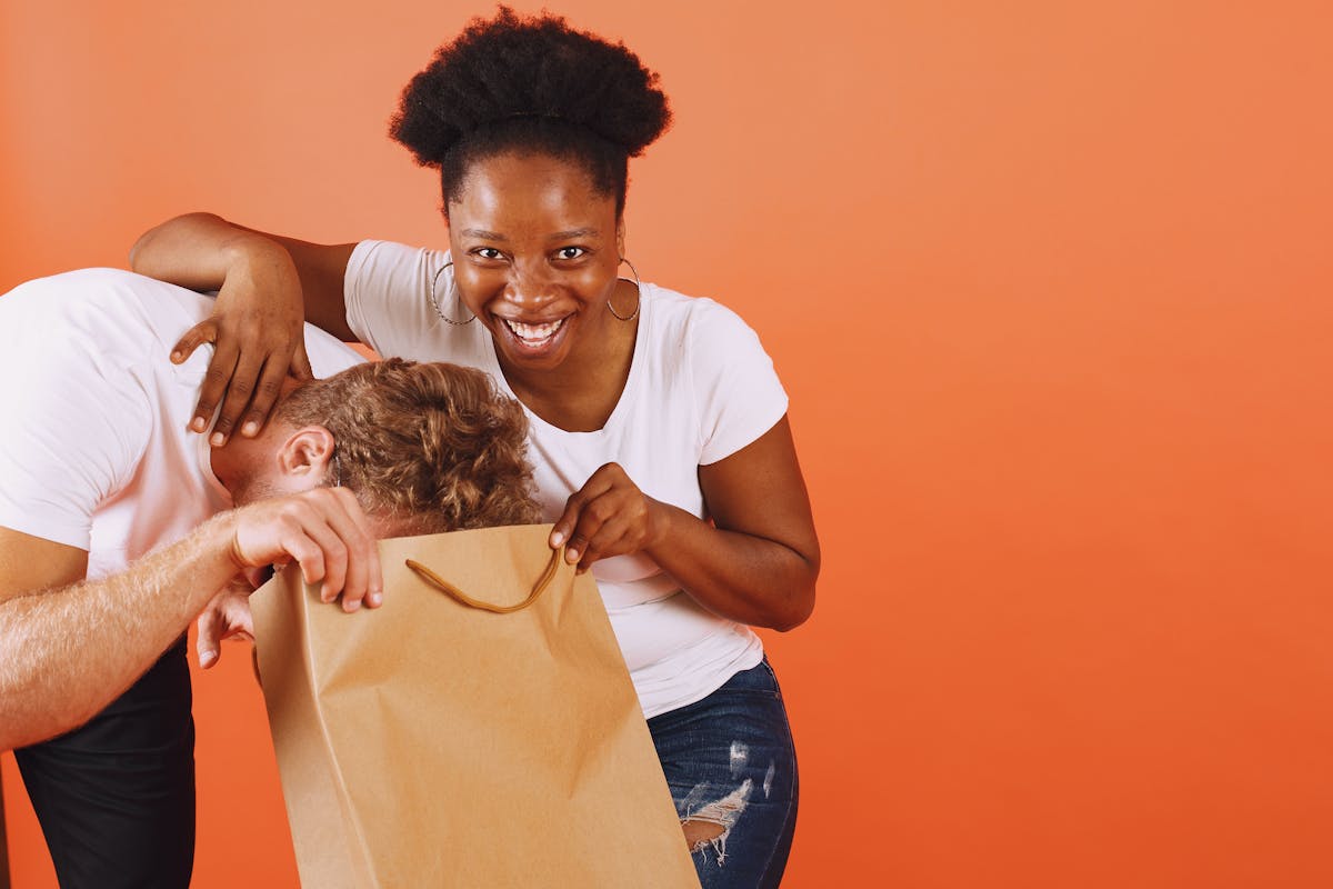 10 Odd Habits That Make Relationships Stronger 3 A happy woman and man having fun with a paper bag against an orange studio backdrop.