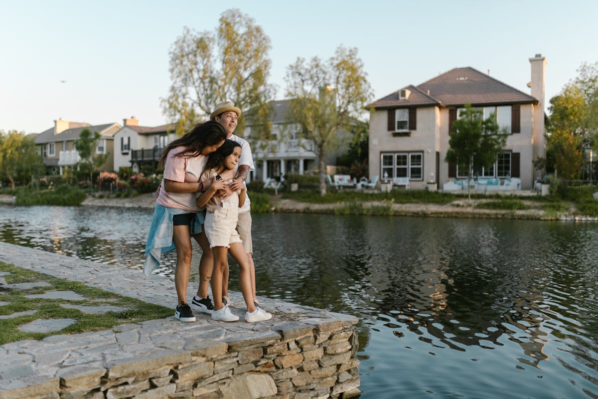 10 Expensive Mistakes to Avoid When Buying a Home 3 A joyful family embraces by a tranquil lake with beautiful houses in the background.