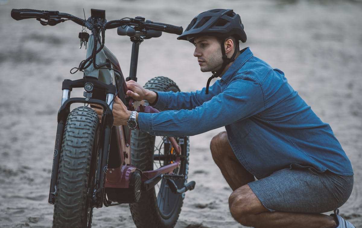 8 Mistakes to Avoid When Purchasing an E-Bike 3 A man in a blue shirt and helmet fixing an electric bicycle on sandy terrain.