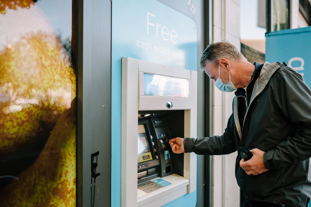 10 Everyday Habits That Are Draining Your Wallet 3 A man wearing a mask using an ATM machine outdoors for cash withdrawal.