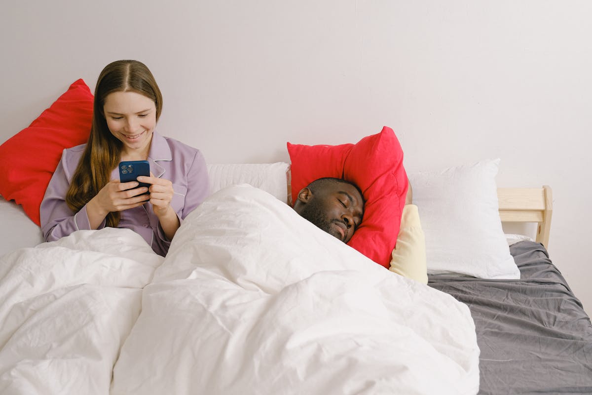 10 Critical Signs Your Marriage Is Heading for Divorce 4 A smiling woman on her phone next to a sleeping man in a cozy bedroom setting.