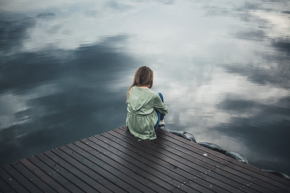 10 Dark Truths You Won't Know About Divorce Until You Get Divorced 4 A solitary woman in a green hoodie sits on a wooden dock, reflecting silently by a placid lake.