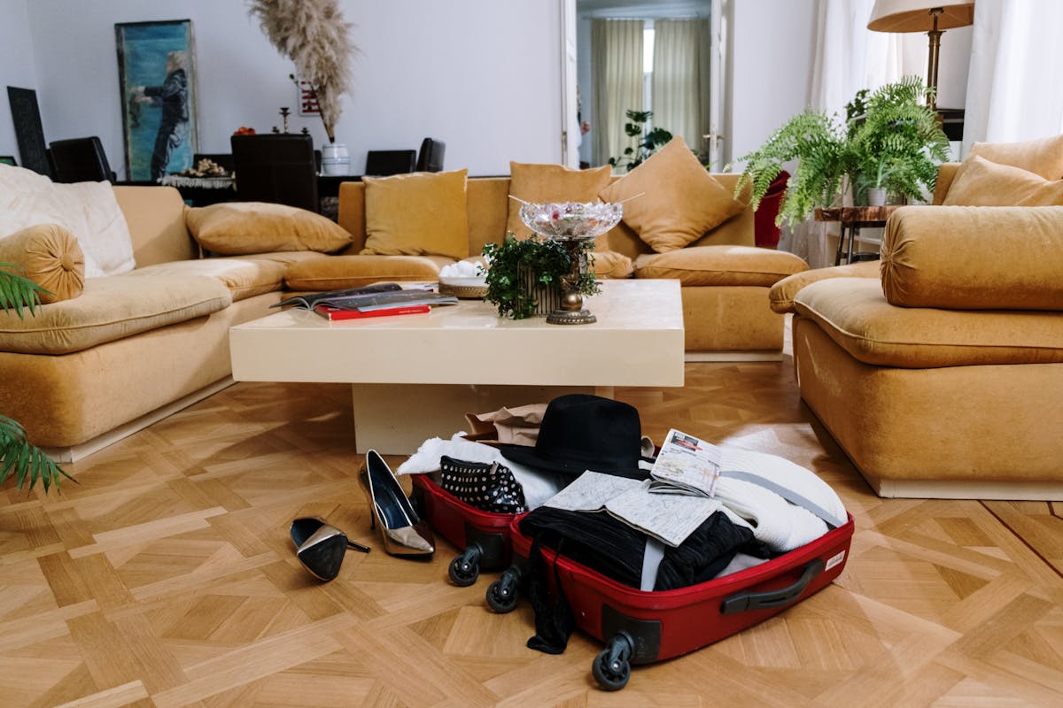 14 Common Travel Mistakes That Will Destroy Your Budget 4 A stylish living room with beige couches and an open suitcase on a parquet floor, surrounded by plants.