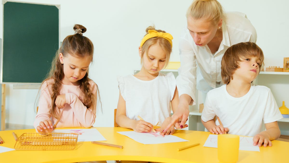 You’re Probably Broke Because of These 6 Hidden Financial Pressures 3 A teacher helping young children with drawing activities in a bright classroom setting.