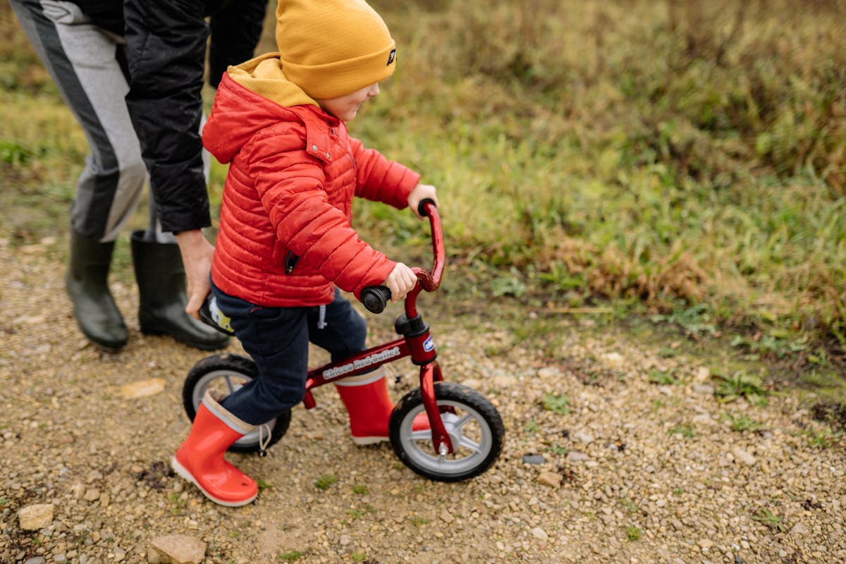 10 Parenting Habits That Drain Your Budget 2 A young child in a red jacket rides a balance bike on an unpaved path outdoors.
