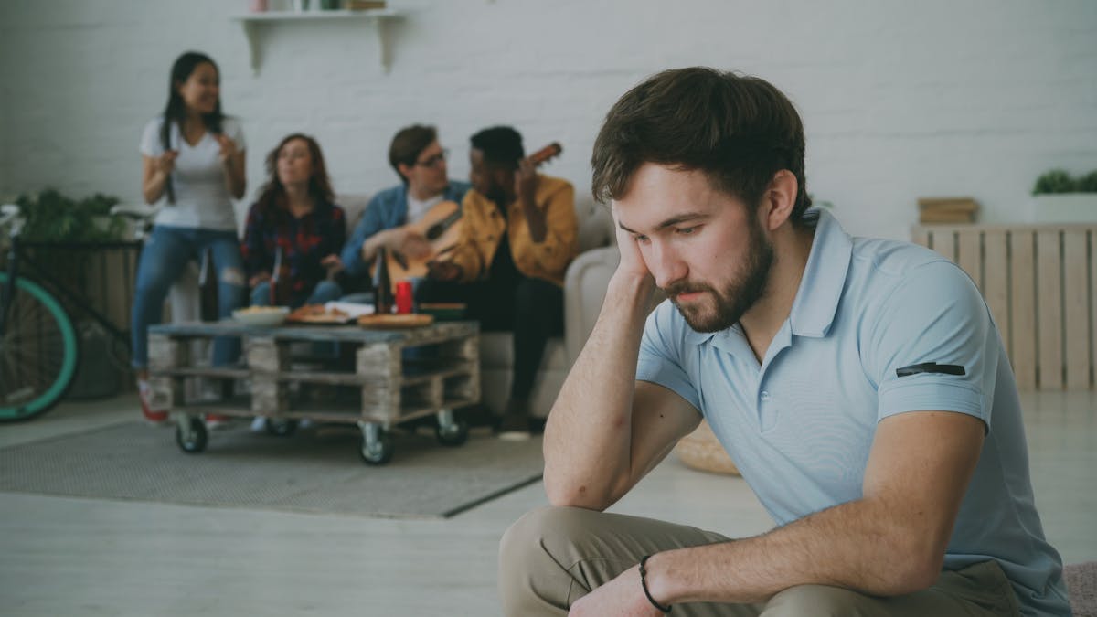8 Signs Your Spouse is Way More Self-Centered Than They Seem 1 A young man sits alone looking somber while friends socialize in the background.