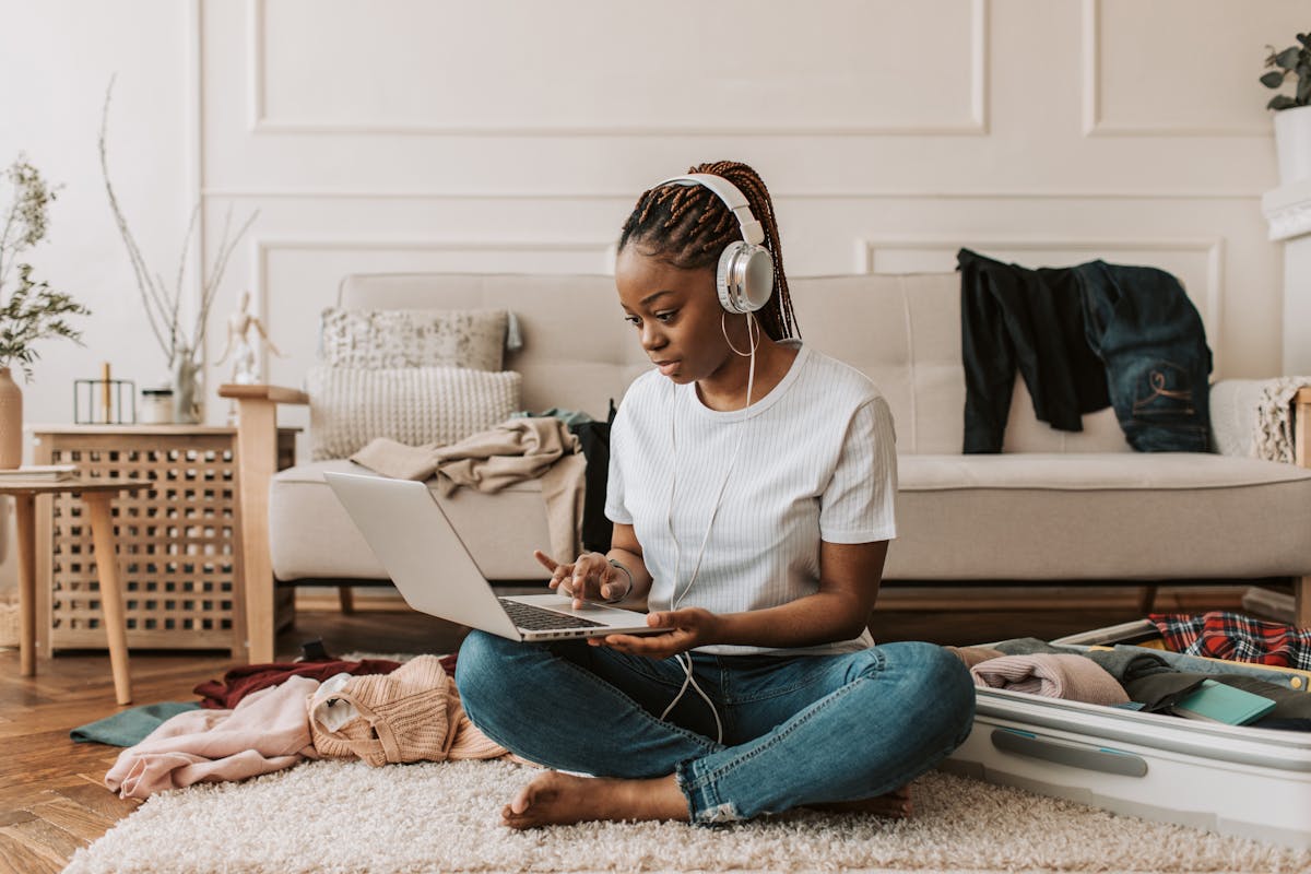 8 Disturbing Secrets Airlines Don’t Want You to Know 2 A young woman sits on the floor using a laptop and headphones, amidst clothes and a suitcase.