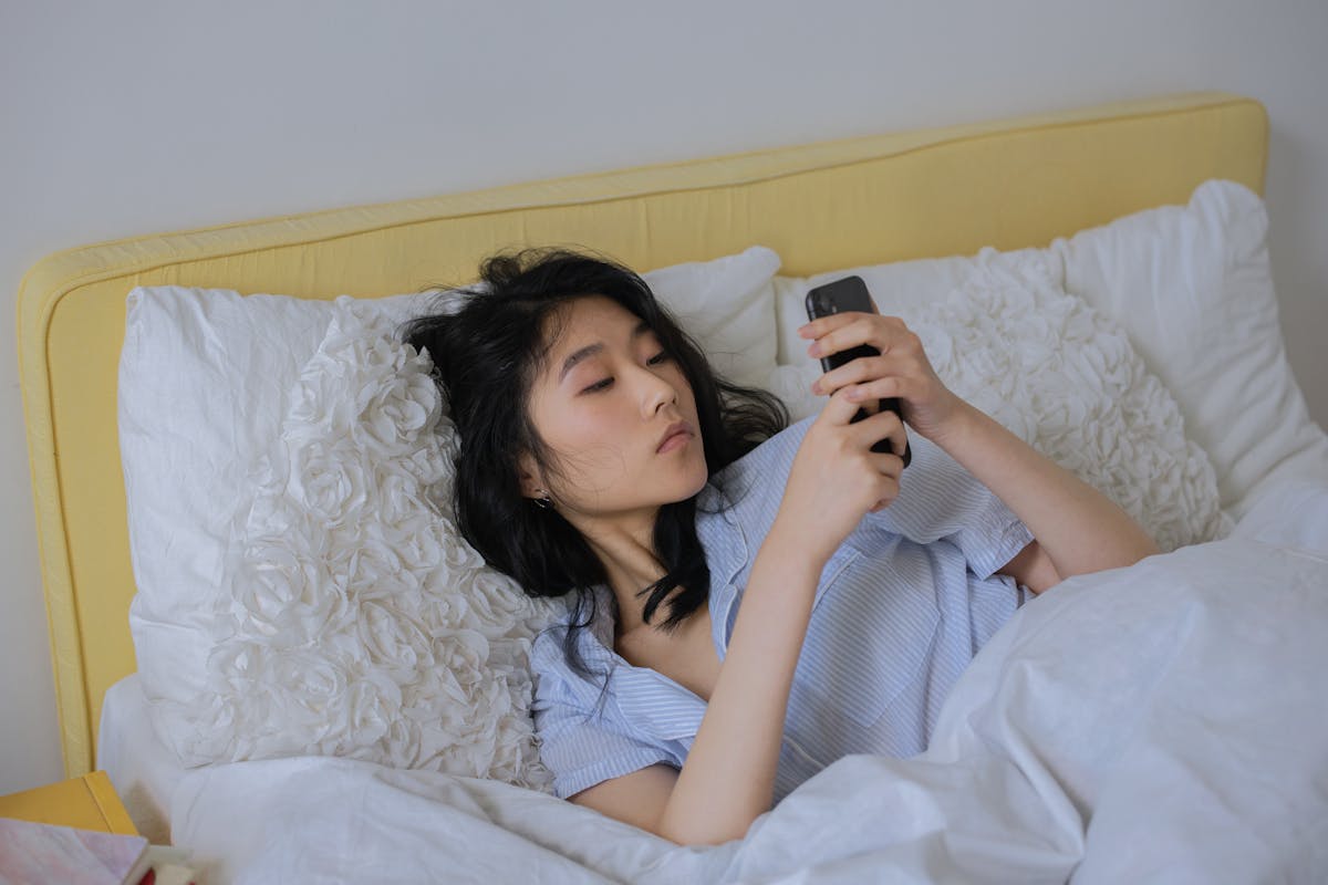 10 Unhealthy Habits That Are Undermining Your Well-being 2 Asian woman comfortably lying in bed, using her phone in a serene bedroom setting.