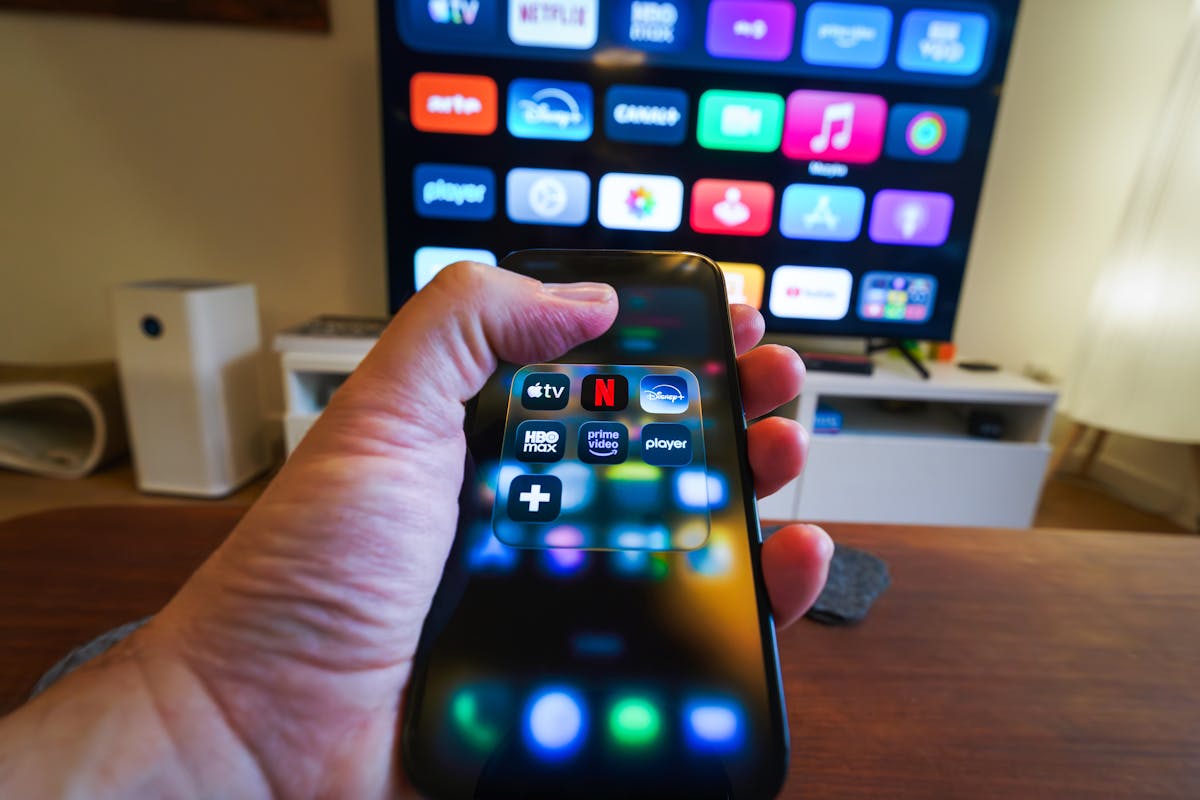 8 Costly Mistakes That Are Quietly Destroying Your Finances 1 Close-up of a hand holding a phone displaying streaming apps in front of a TV with multiple app icons.