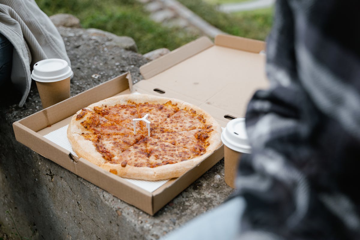 8 Worst Foods to Eat Before Bed That Quietly Destroy a Good Night’s Sleep 1 Close-up of a pizza in a box with coffee cups on a stone surface outdoors.