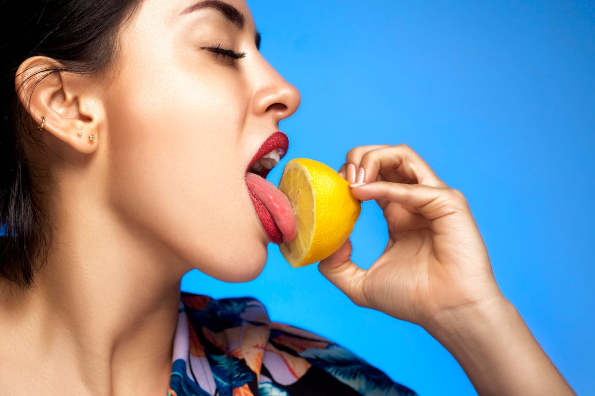 8 Odd Health Symptoms We Should Never Ignore 4 Close-up of a woman licking a lemon slice with a vibrant blue backdrop. Fresh, vibrant, and bold imagery.