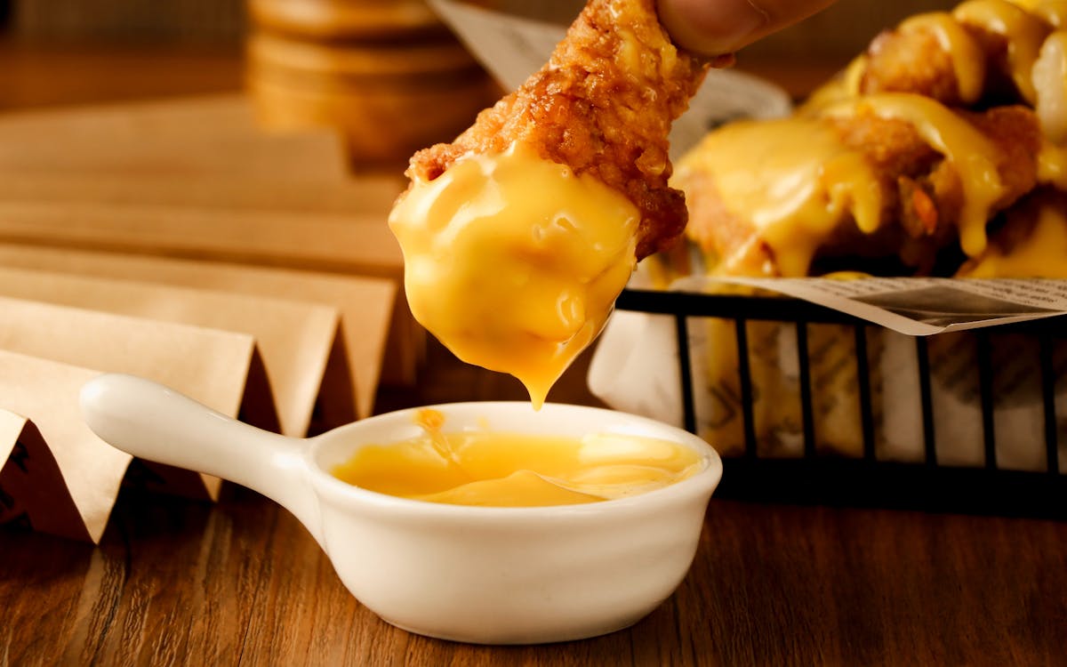 9 Surprising Facts About American Cheese That Will Blow Your Mind 2 Close-up of crispy chicken tenders being dipped into a creamy cheese sauce.