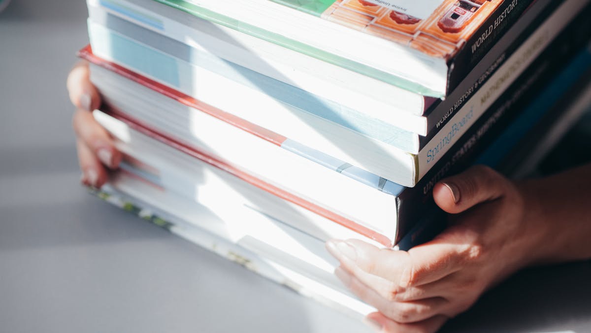 10 Things to Never Pay Full Price For 3 Close-up of hands holding a stack of colorful books in natural light.