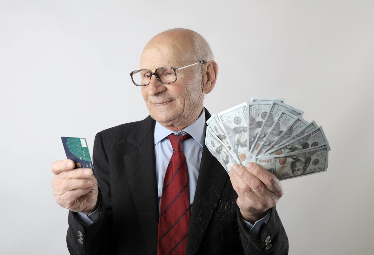 10 Money Mistakes That Are Keeping You Broke And Frustrated 3 Elderly man in a suit holding a credit card and US dollar bills, representing finance and wealth.