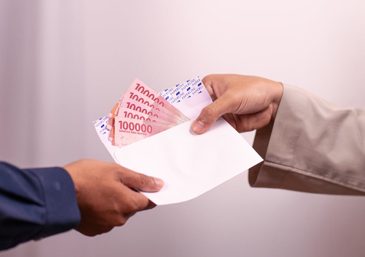 10 Bizarre Schemes Corporations Have Tried To Get Away With 3 Hands giving and receiving Indonesian rupiah in an envelope, symbolizing financial transaction.