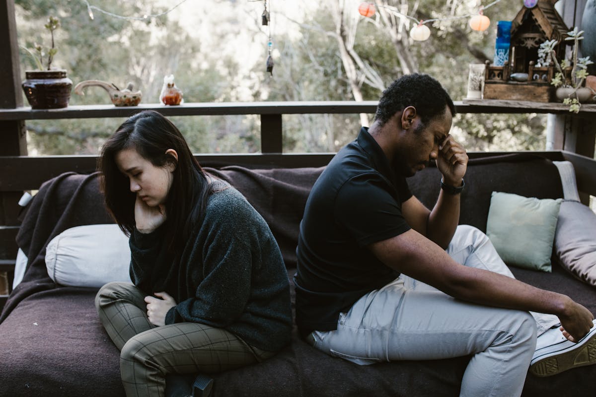 If You Ignore These 8 Red Flags, Your Boyfriend Will Be a Dud in a Year 2 Interracial couple sitting back to back, reflecting emotional conflict, outdoors.