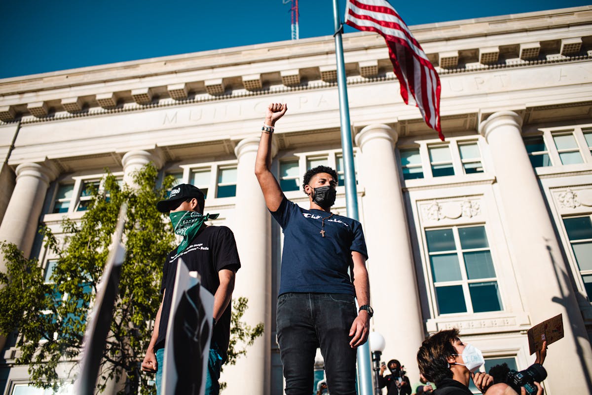 10 Political Scandals That Shook Nations and Triggered Mass Uprisings 4 Protesters in Des Moines advocate for freedom with raised fists and American flag backdrop.