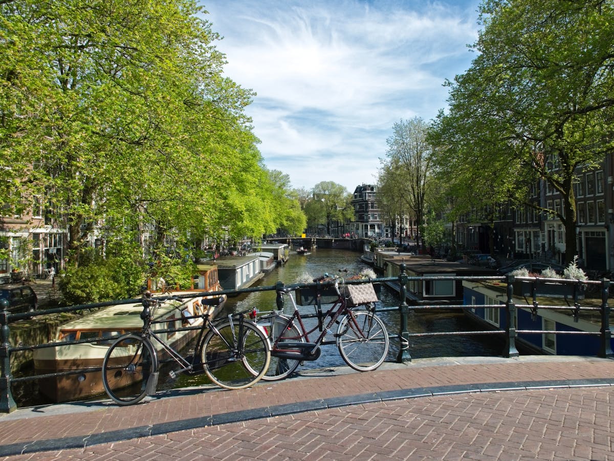 7 Overcrowded Destinations to Skip This Summer 3 Scenic view of bicycles by a canal in Amsterdam with springtime greenery and blue skies.