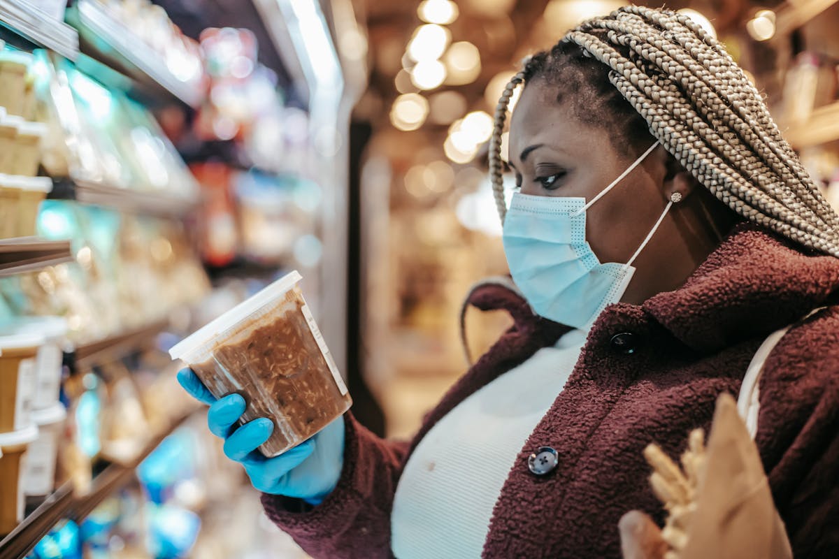 8 Secrets About the Food Industry They Don’t Want You to Know 2 Side view of African American female customer on medical mask reading label on frozen food while shopping in grocery store
