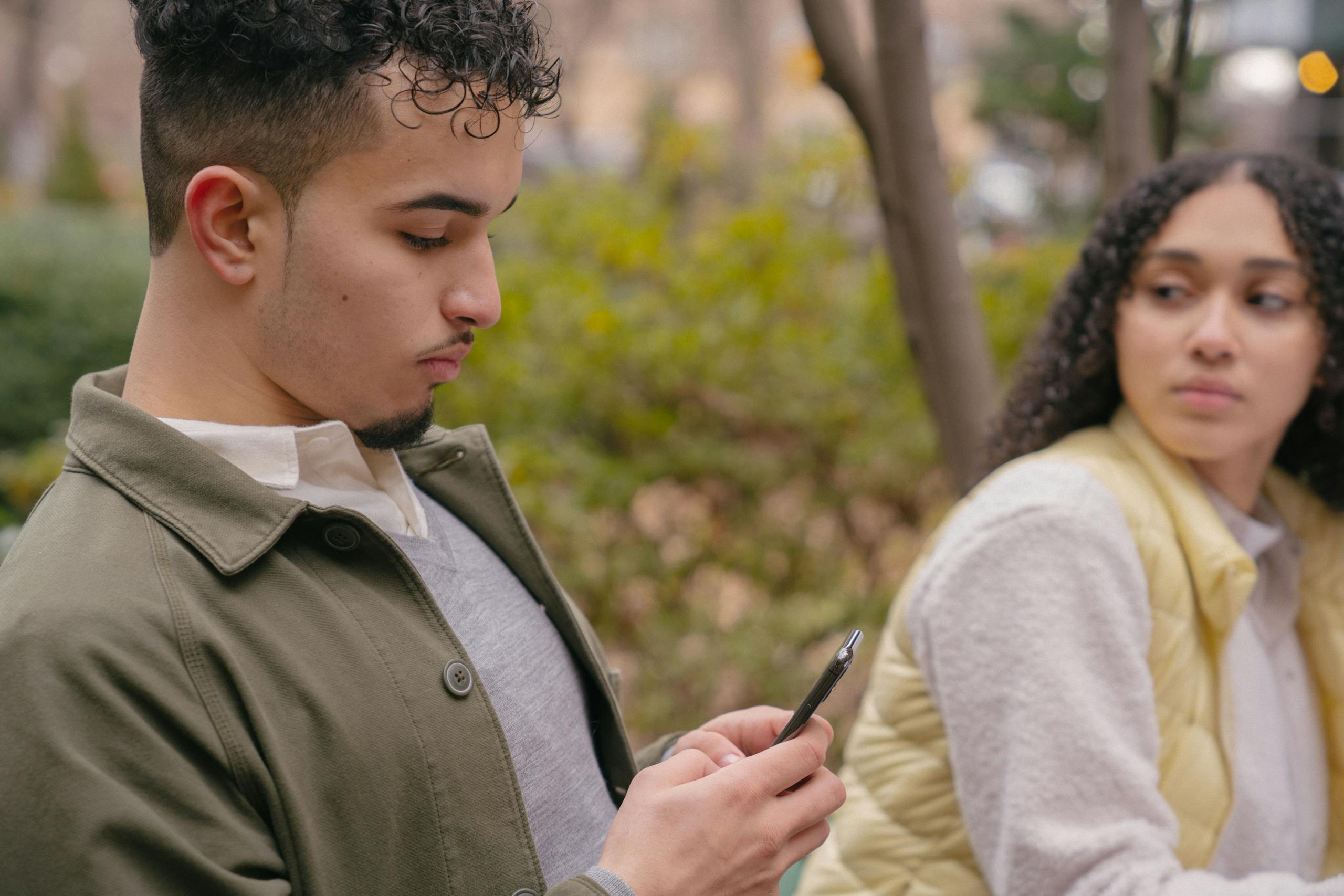 Side view of Latin American male surfing on phone while girlfriend looking seriously and sitting in park near trees and bushes in daytime