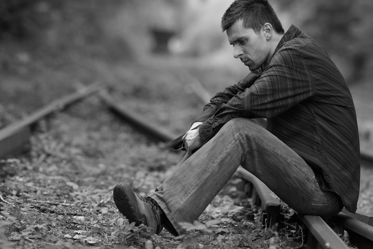 12 Reasons to Seek Therapy 4 Thoughtful man sitting on abandoned railroad tracks, evoking solitude and contemplation in black and white.