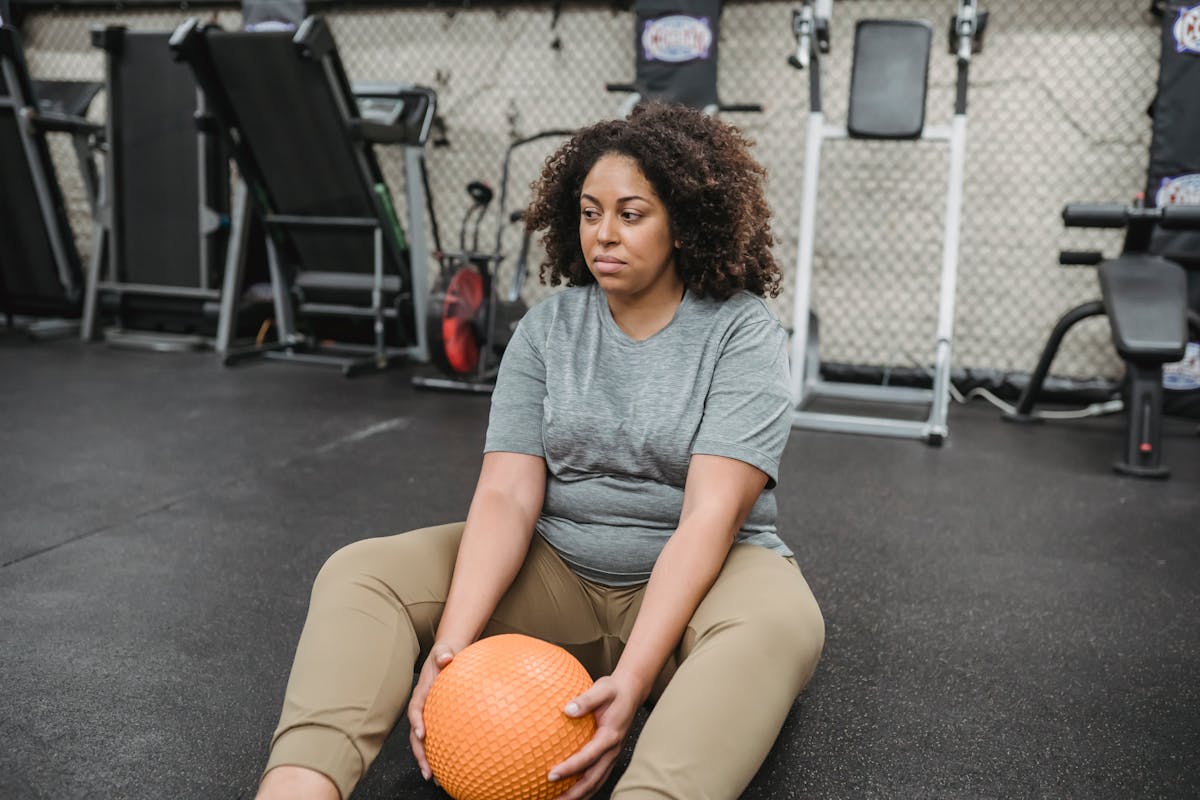 8 Motivation Mistakes That Are Killing Your Workout Routine 1 Tired plus size young African American female with curly hair in activewear sitting on floor with medicine ball after abs workout in gym