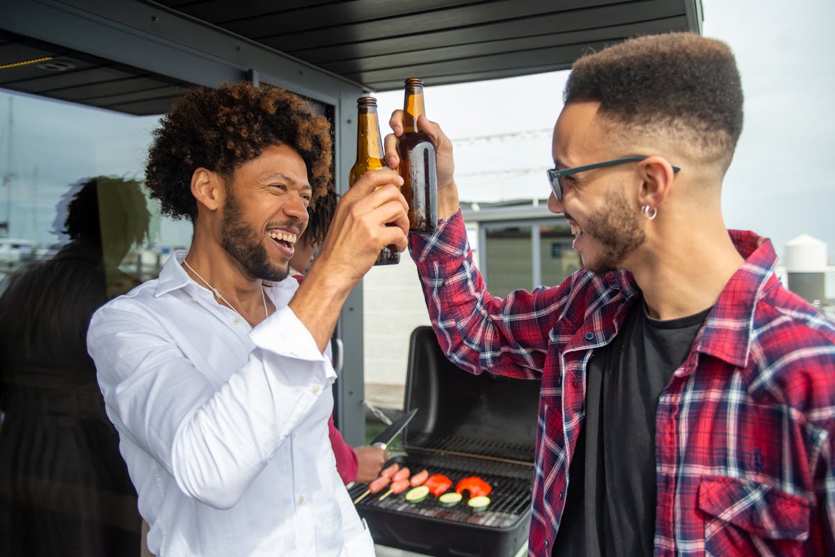 10 Cheap and Fun Bachelor Party Ideas 1 Two friends clink beer bottles while cooking at an outdoor barbecue in Portugal.