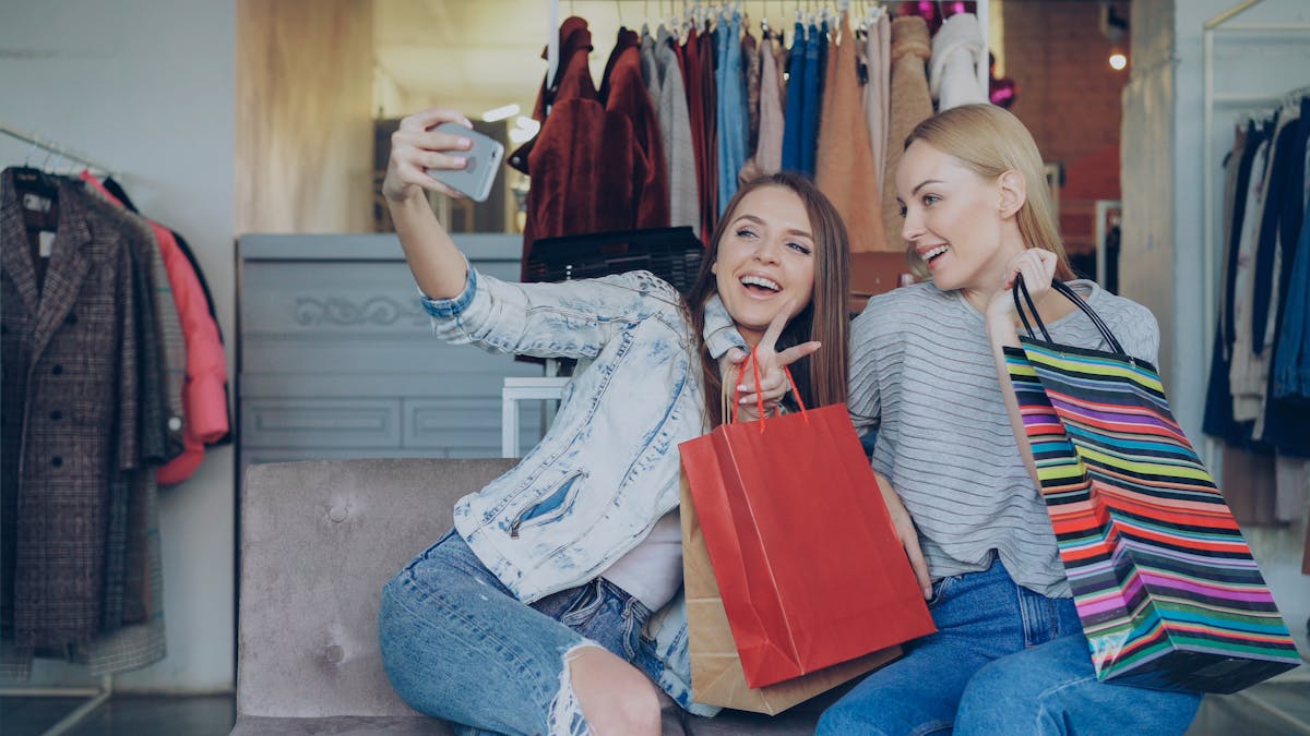 Ignoring These 6 Financial Mistakes Could Cost You Everything 2 Two young women enjoying shopping, taking a selfie in a stylish boutique with shopping bags.