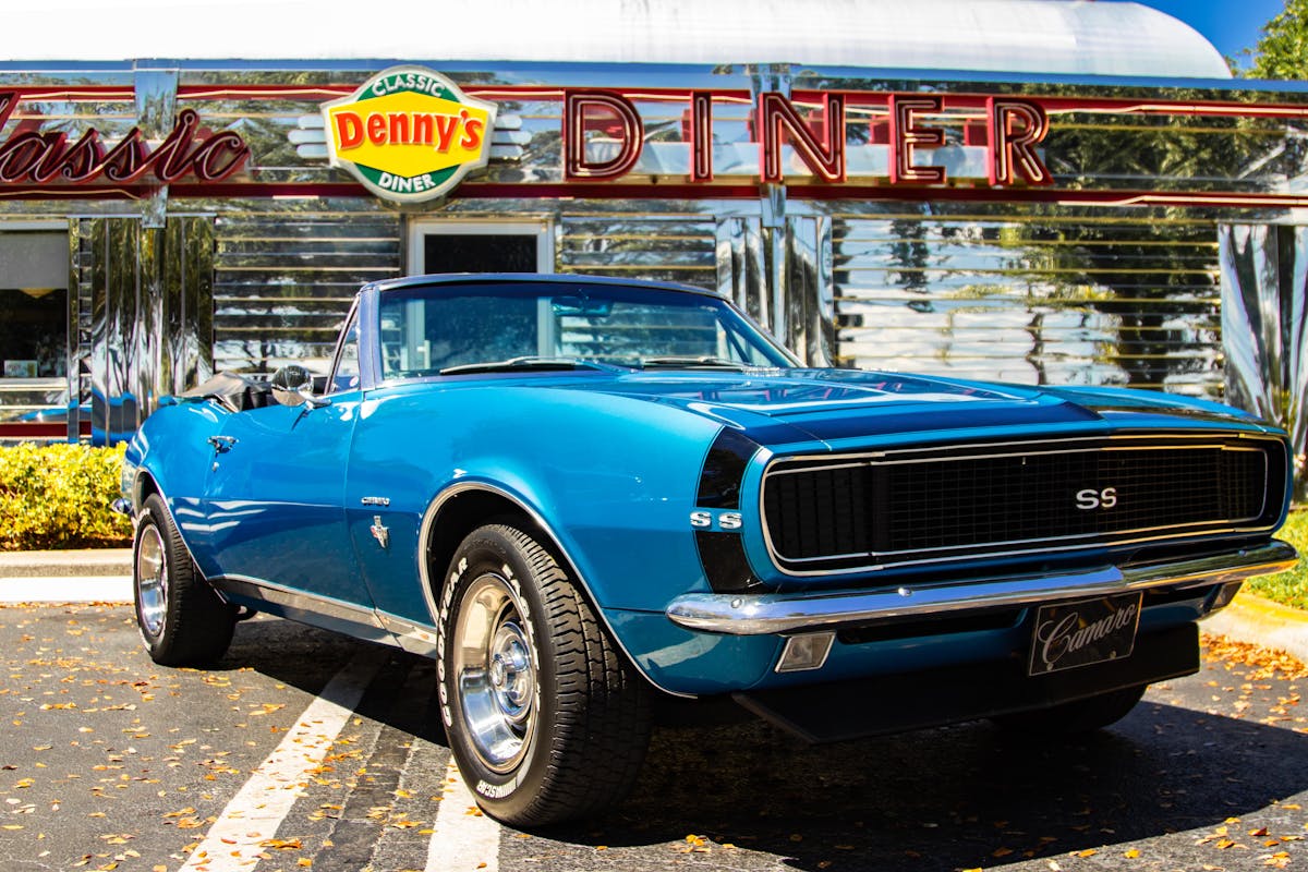 8 Overrated Chain Restaurants That No Longer Earn Their Price Tag 4 Vintage blue Chevrolet Camaro parked in front of Denny's Classic Diner under sunny skies.