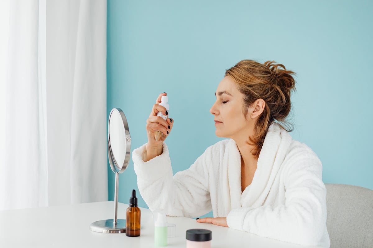 9 Mistakes to Avoid When Preparing for Summer 2 Woman in a bathrobe using a skincare spray in front of a mirror indoors.