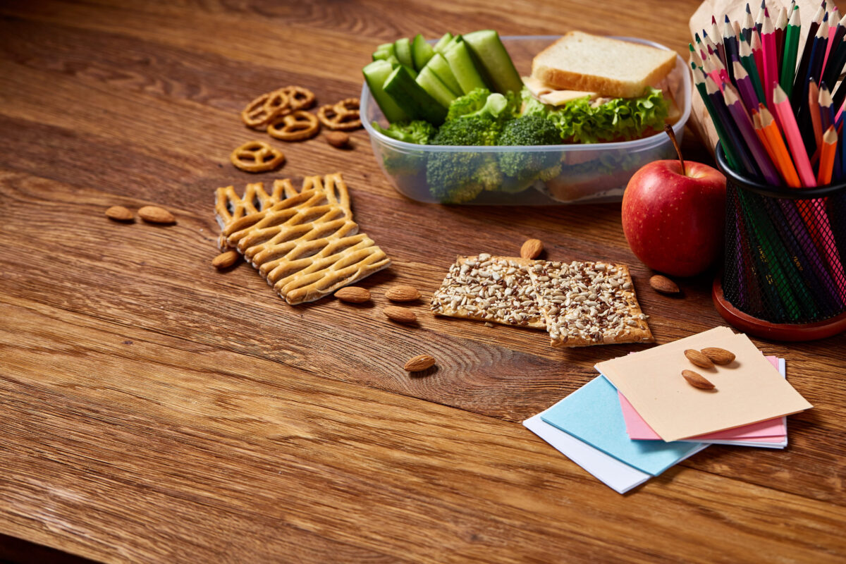12 School Lunches You Should Stop Packing For Your Kids Immediately 1 Tasty lunch with green lettuce and fresh vegetables and apple in transparent plastic container and different school supplies on wooden desk, selective focus, close-up. School lunch concept. Healthy eating concept.