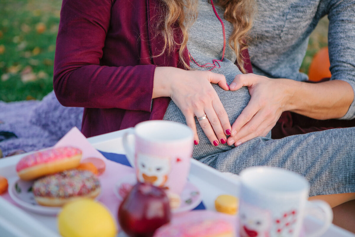Male and female hand of spouses with their palms made a heart shape on the round belly of a pregnant wife on a blurred background picnic in nature, selective focus