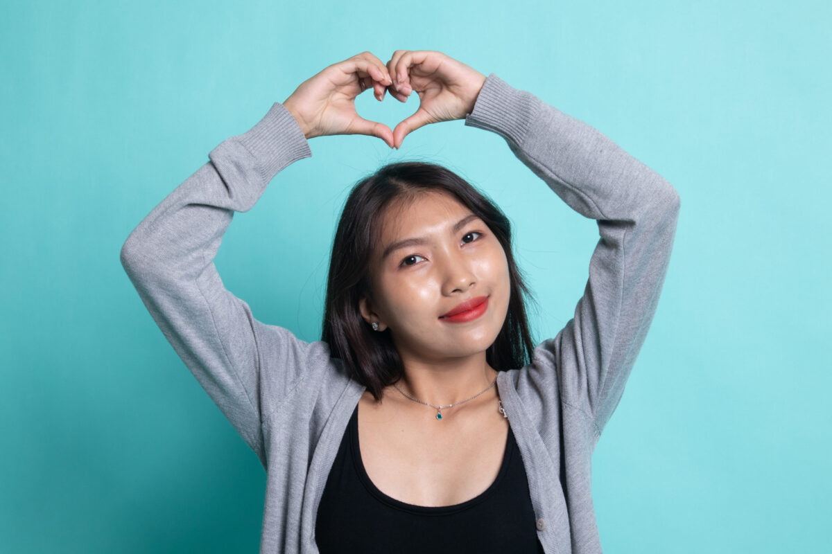 10 Reasons Why Many Relationships Survive on How Much a Woman Is Expected to Suffer 3 Young Asian woman gesturing heart hand sign on cyan background.