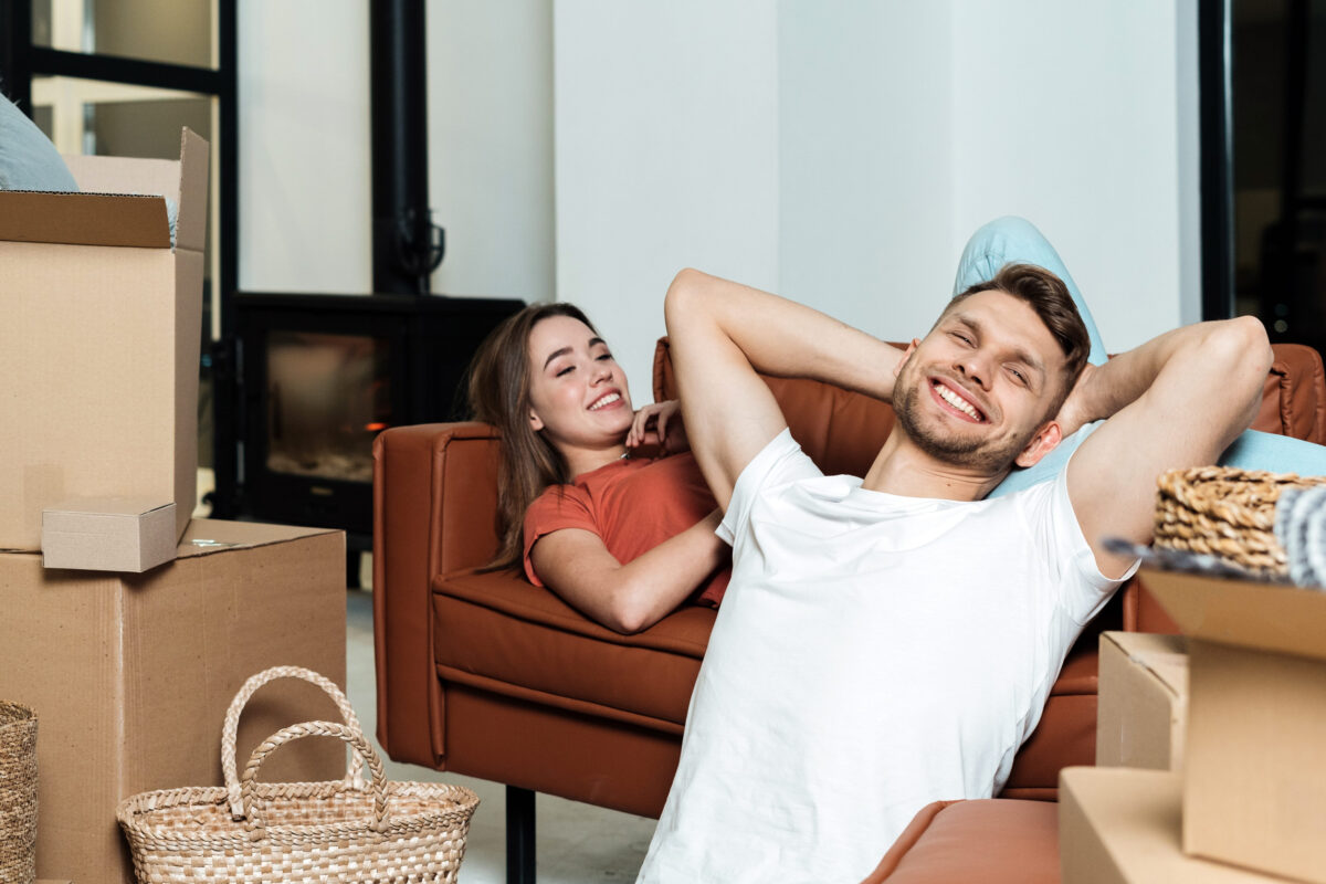 Happy and dreamy young adult couple move in new house together, smiling wide, making cheerful faces, resting in living room after unpacking carton boxes